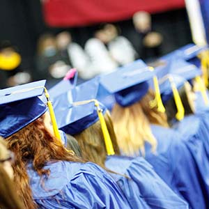 A line of graduates in blue caps and gowns march in a procession towards a stage.