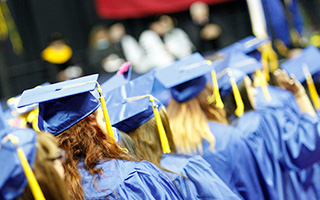 A crowd of graduates clad in red caps and gowns. One grinning graduate stands tall above the others with their hands held high above their head.